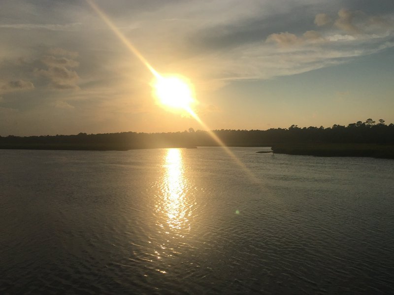Bright golden sunset casts a sparkling reflection across calm rippled river water lined with tree...