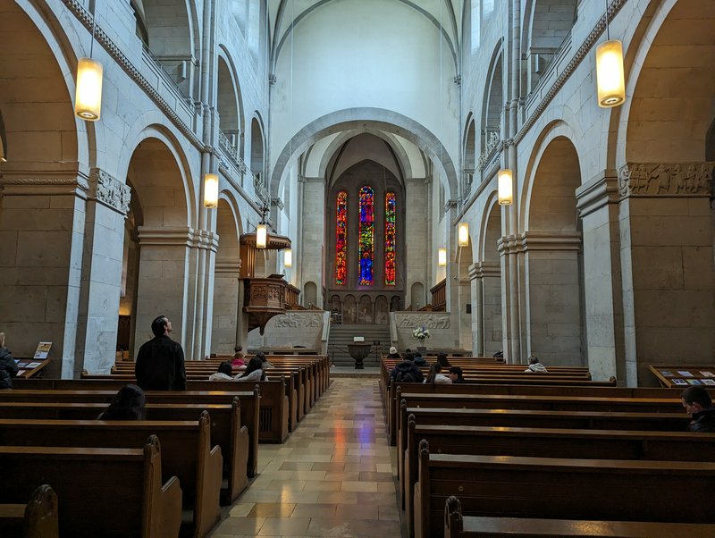 Stone cathedral nave with wooden pews, tall stained glass windows, and quiet visiting congregants