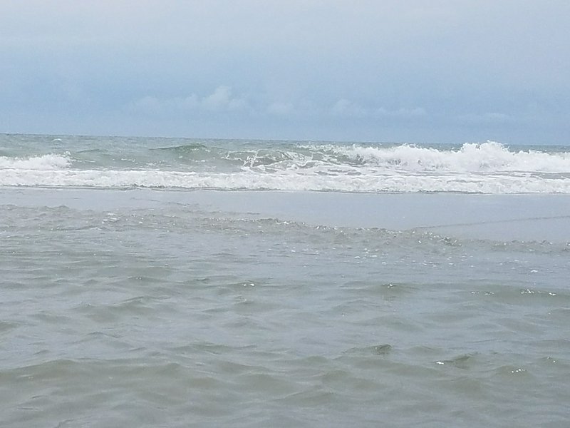 Rolling foamy ocean waves approach a shoreline under a pale overcast blue sky