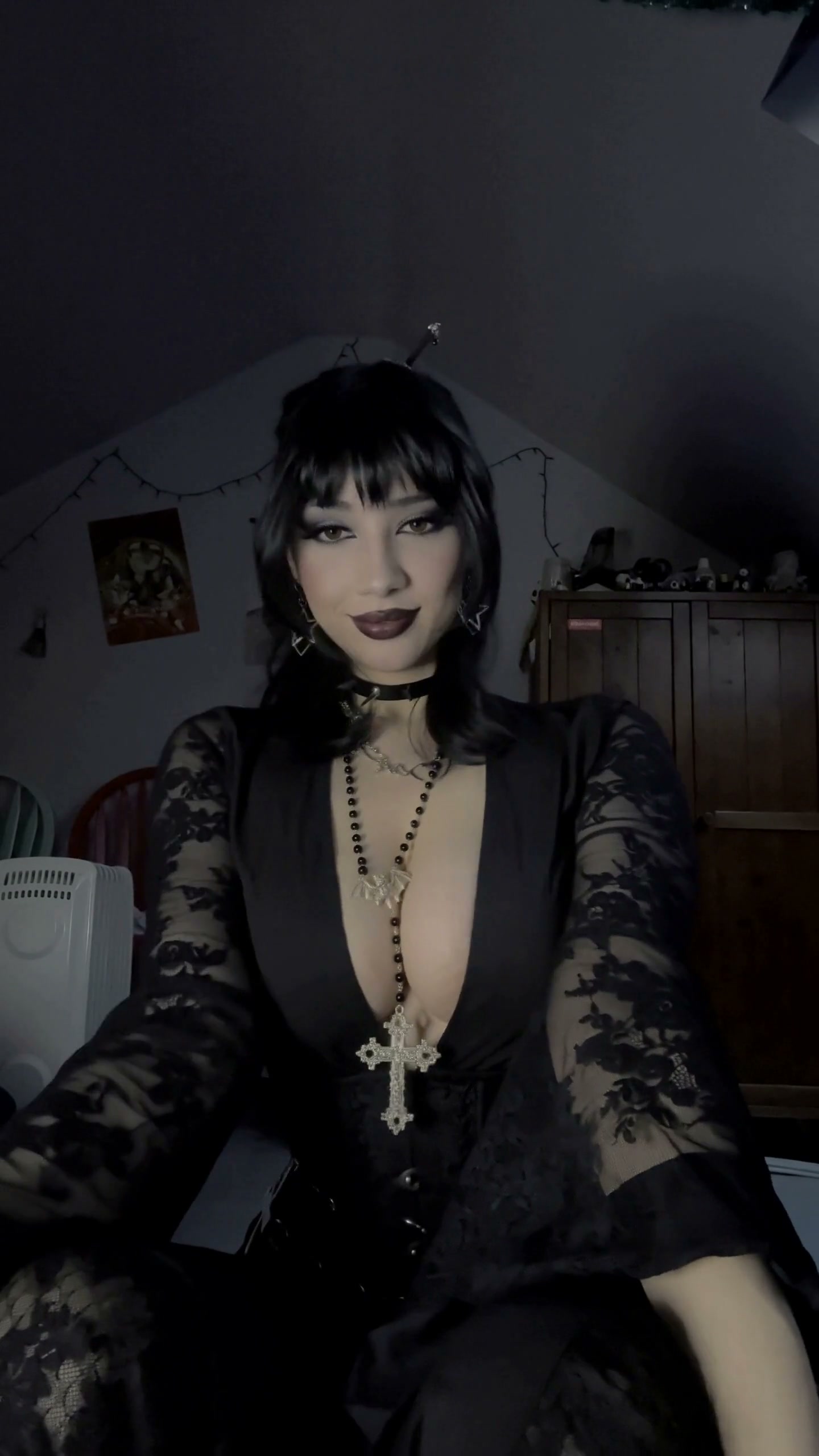 Gothic styled woman in black lace outfit with ornate cross rosary, posing in a dim cozy attic bed...