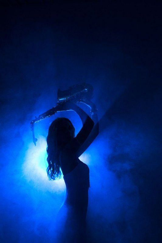 Silhouette of a female musician holding a saxophone above her head against glowing smoky blue sta...