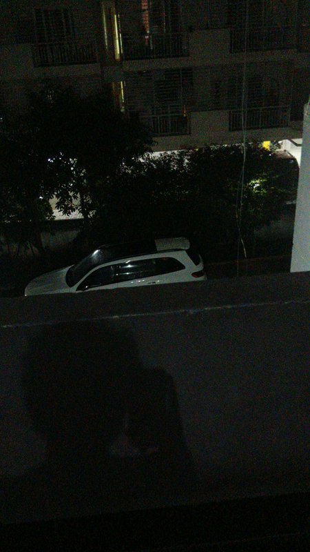 Night view from a residential balcony showing a parked white SUV, leafy trees, and an apartment b...