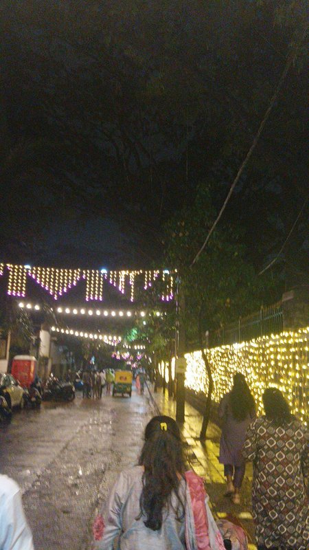 Rain-soaked nighttime street strung with festive fairy lights, pedestrians walking past lit fenci...