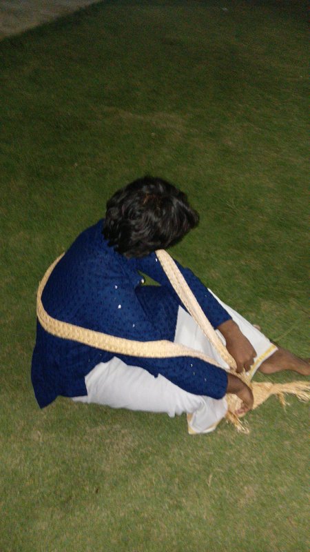 Person in traditional South Asian clothing sits on grass holding a thick woven ceremonial straw rope
