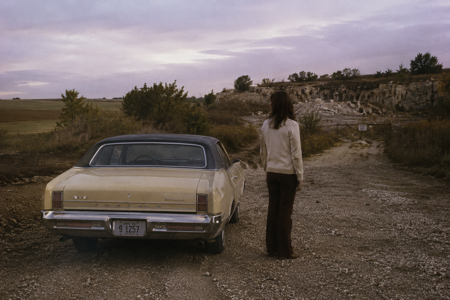 Vintage beige car parked on a gravel road as a woman stands nearby in a rural landscape.