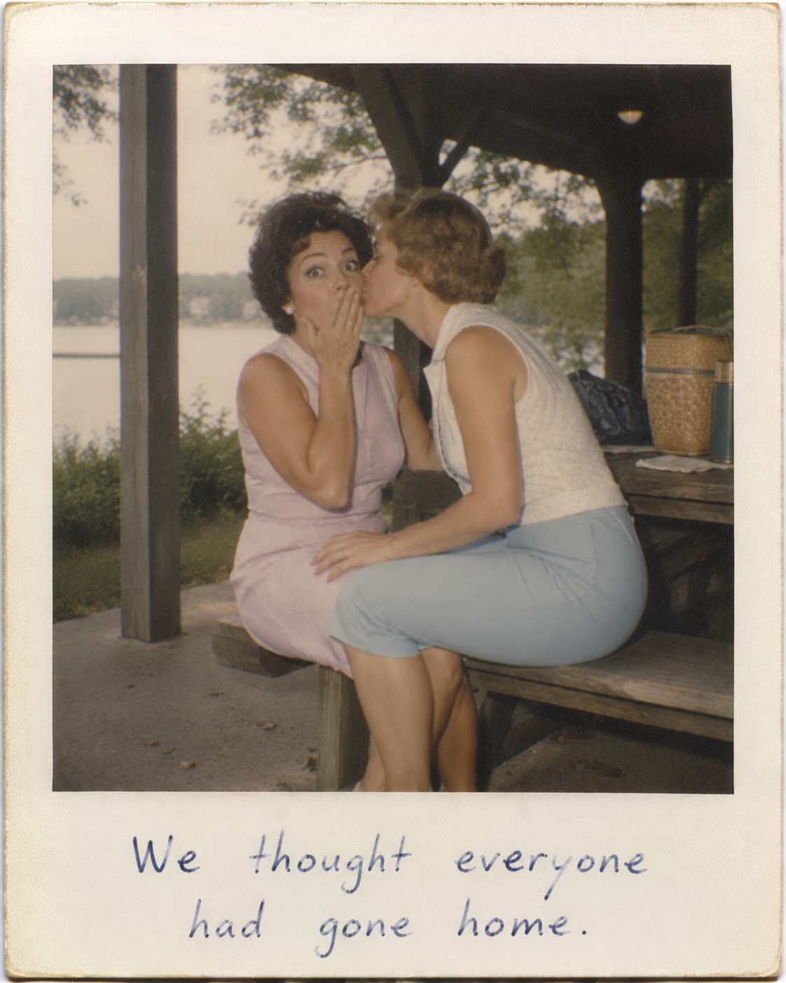 Two women sharing a kiss on a park bench under a gazebo by a lake in a vintage photo.