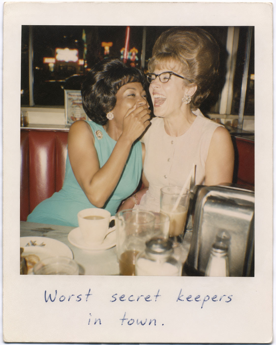 Two women laughing together in a retro diner, one whispering a secret.