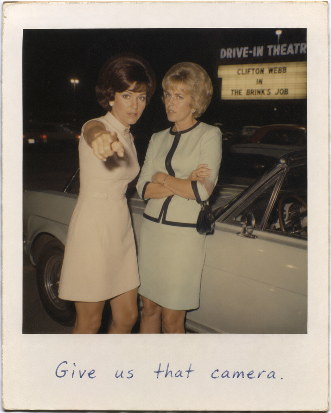 Two women in 1960s dresses posing beside a white vintage car at a drive-in theater, one pointing at the camera.
