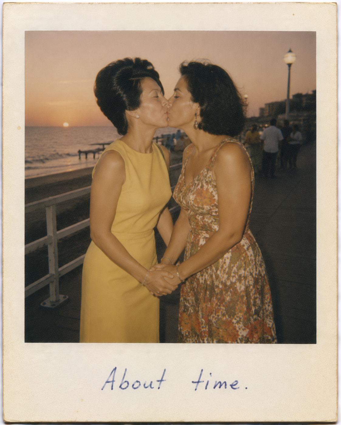 Two women kiss on a seaside boardwalk at sunset, holding hands.