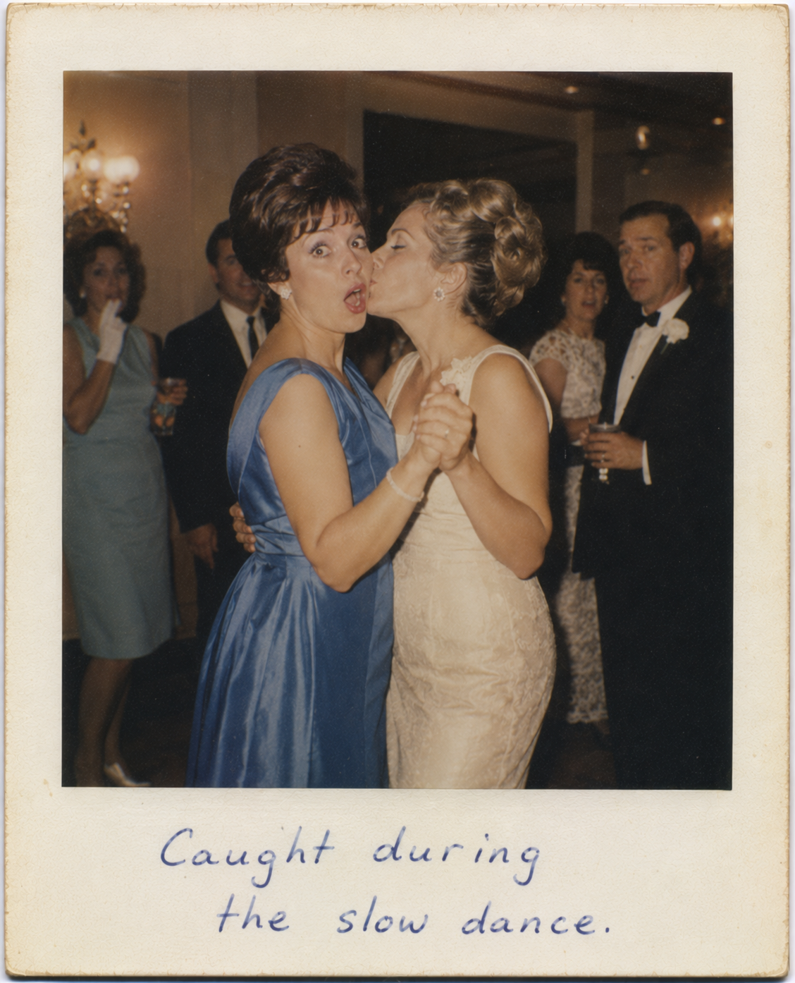 Two women in formal dresses share a slow dance at a wedding reception, one leaning in to kiss the other on the cheek.