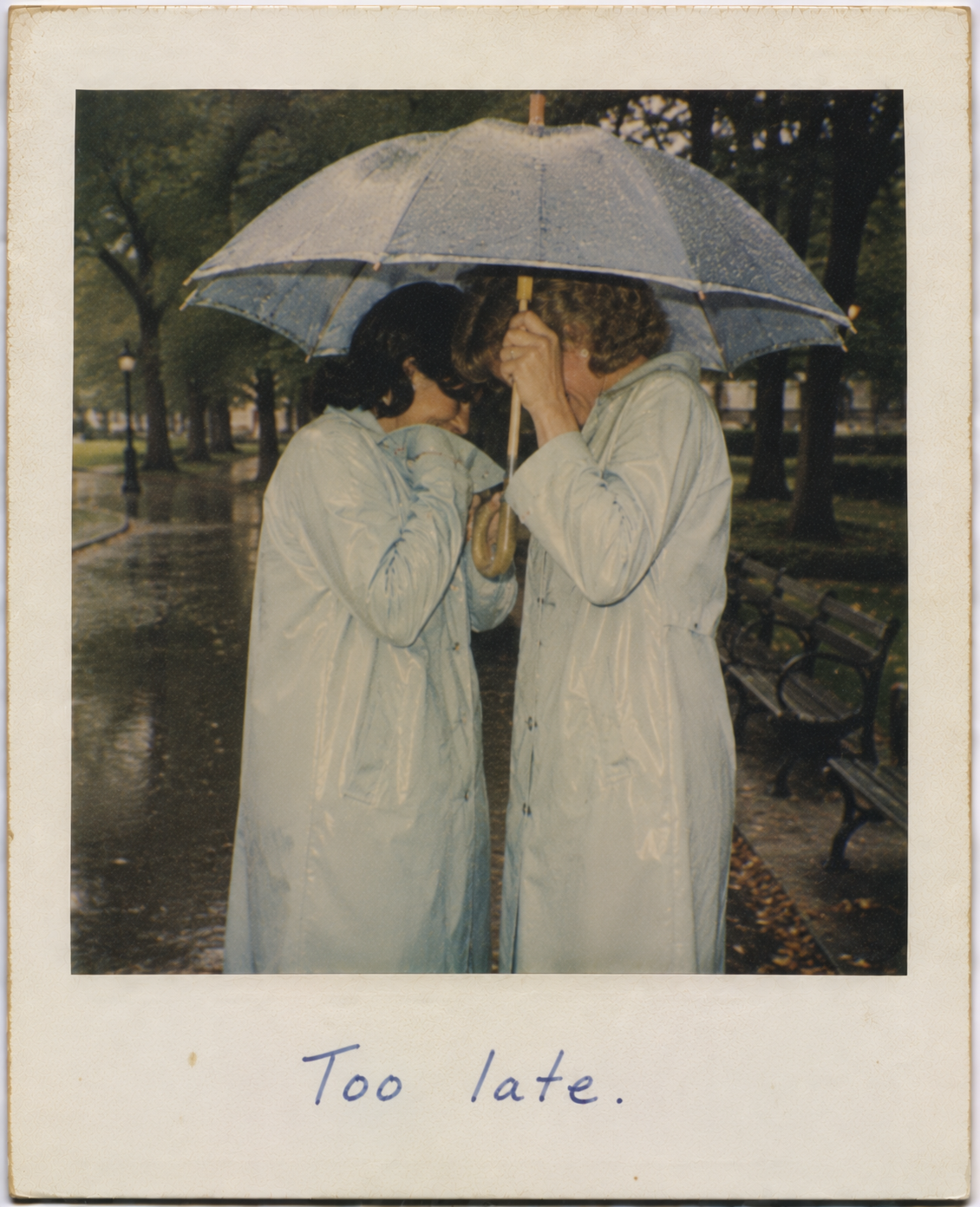 Two people in light blue raincoats sharing a kiss under a large umbrella in a rainy park.