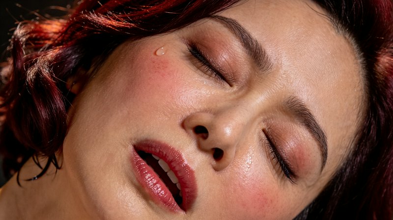 Close-up portrait of a woman with dewy skin, pink lips, and red-toned makeup with eyes closed.