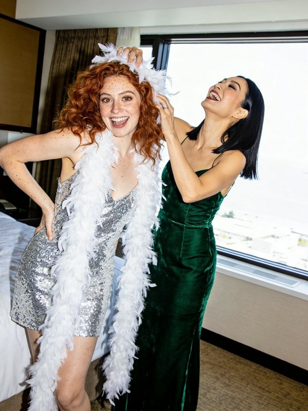 Two joyful women in a hotel room; one with red curly hair in a silver sequin dress and feather boa, the other in a green