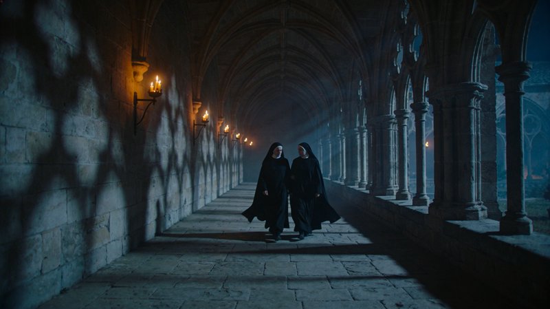 Two nuns walking in a dim, torch-lit medieval cloister with stone arches.