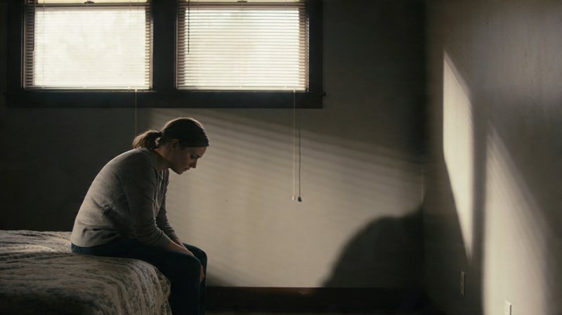 A person sitting on a bed in a dimly lit bedroom with blinds, sunlight streaming across the wall.