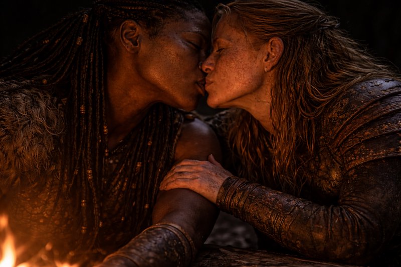 Two women kiss in muddy clothing by a warm campfire.
