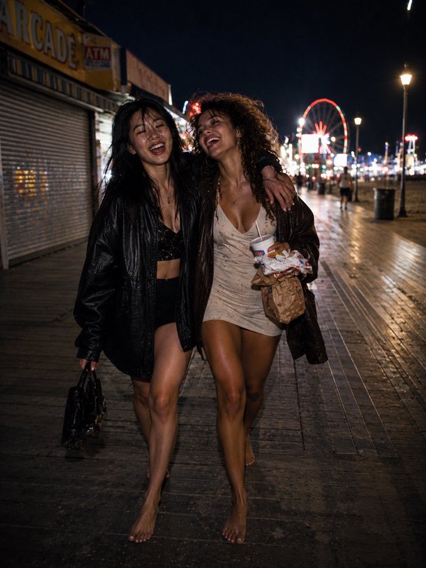 Two young women walking arm-in-arm on a nightboardwalk, smiling and barefoot, under neon lights.