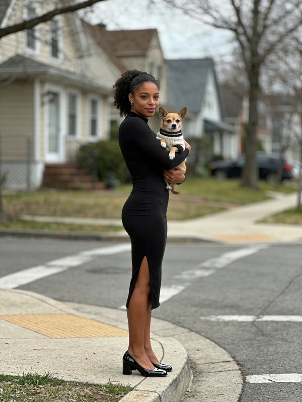 A woman in a black dress holding a small dog on a suburban street.