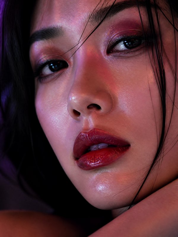 Close-up portrait of a woman with glossy red lips and pink eye makeup under moody lighting.