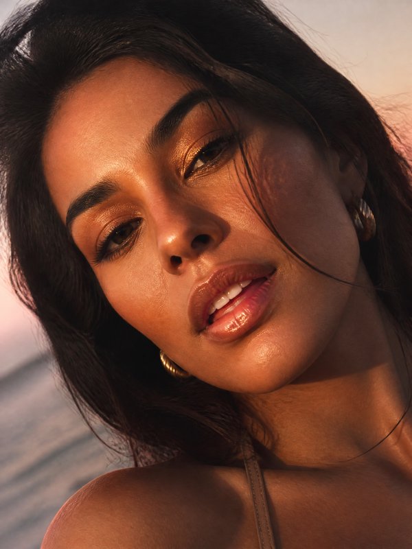 Close-up portrait of a woman at sunset with golden makeup and glossy lips by the beach.