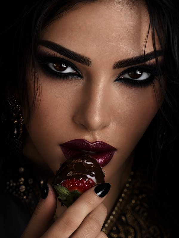 Close-up of a woman with dramatic dark makeup holding a chocolate-covered strawberry.