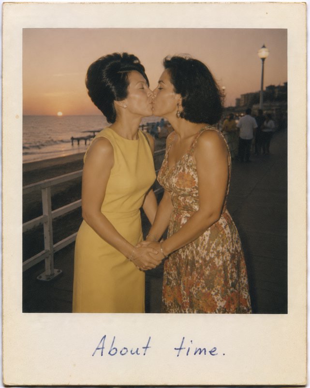 Two women kiss on a seaside boardwalk at sunset, holding hands.