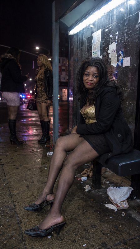 Tired woman sits on a rain-soaked city bus stop bench at night, other women stand on littered wet sidewalk in background.