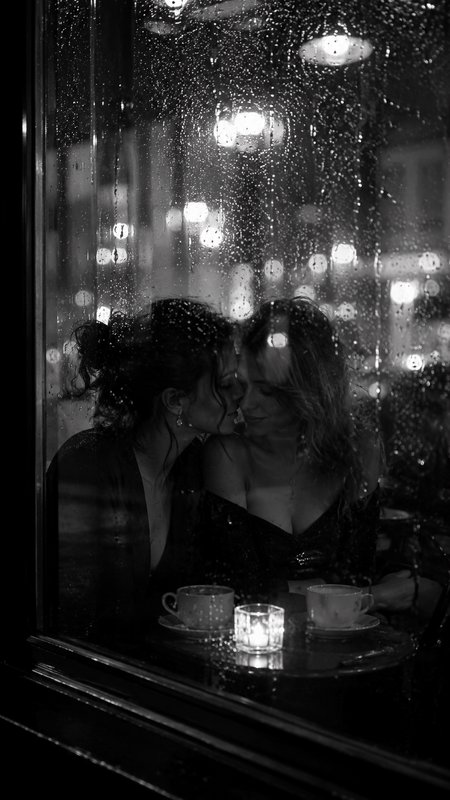 [Black and white intimate portrait of a female couple sharing a tender moment behind a rain-streaked cafe window on a rain...