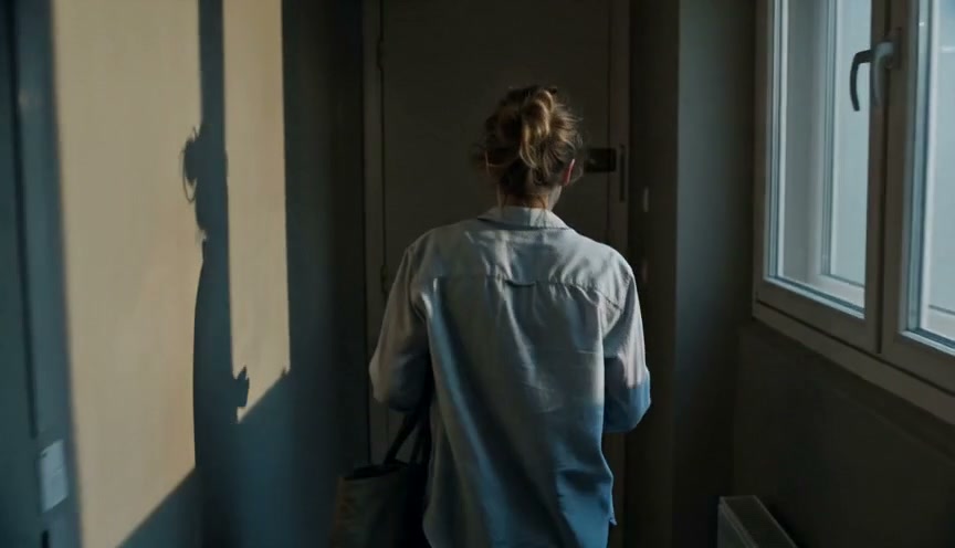 Woman in a light denim shirt walks down a dim hallway, seen from behind, carrying a bag.