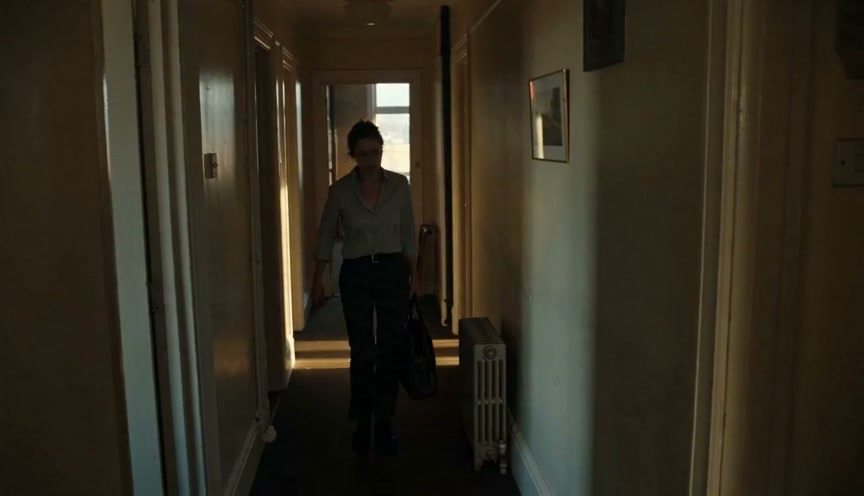 Woman walking down a dim hallway carrying a bag, backlit by a distant light.