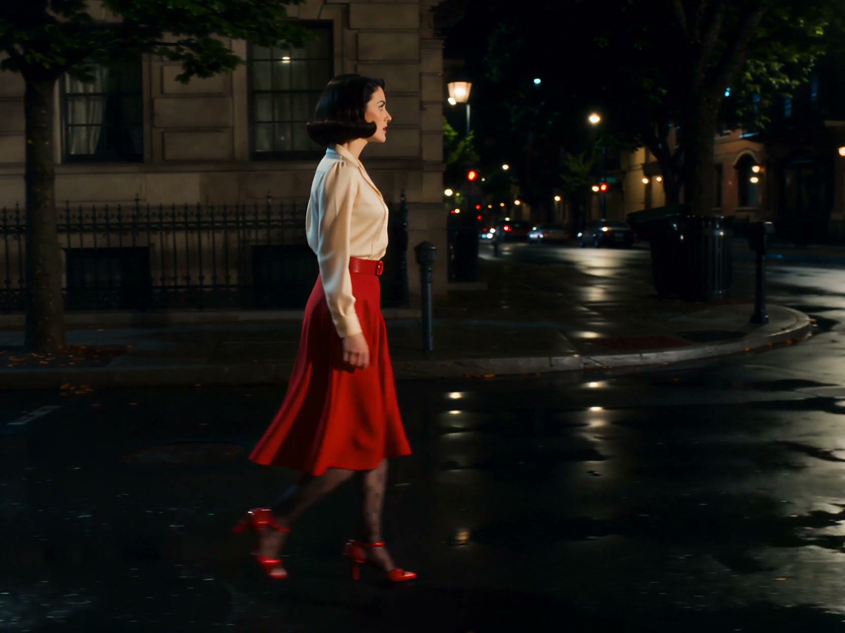Woman in white blouse and red skirt crossing a nighttime, wet city street with reflections.