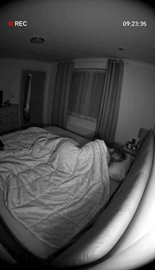 Night vision home security camera view of a person sleeping in a bedroom, with live recording tim...