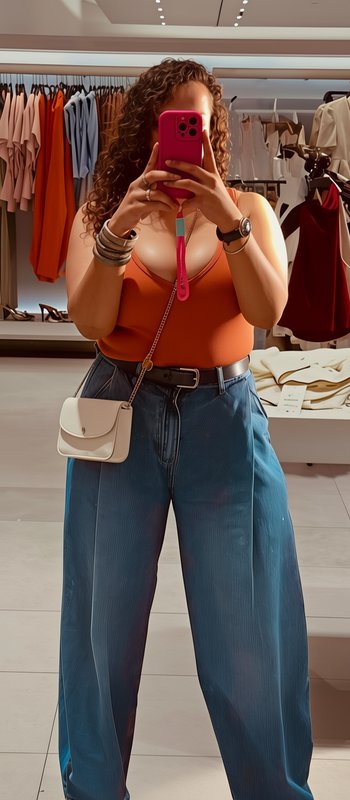Woman takes mirror selfie in a clothing store wearing a rust tank top, wide leg jeans, and small ...