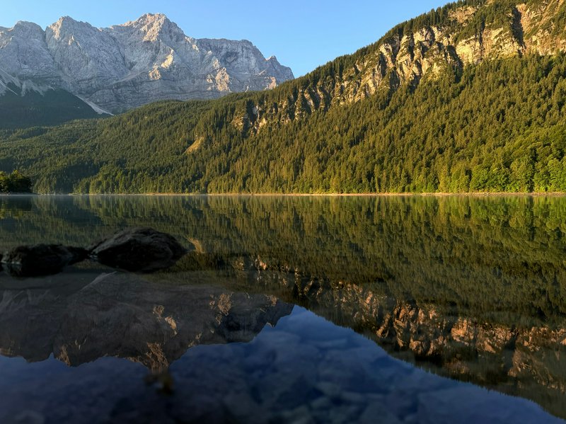Calm crystal alpine lake mirrors forested slopes and snow-capped mountain peaks under a clear sunny blue sky.