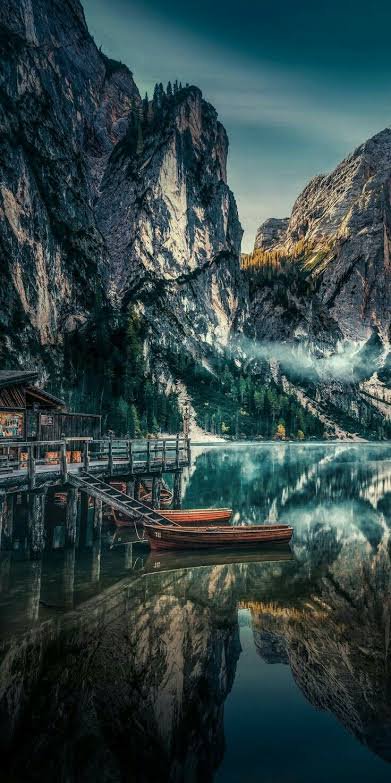 Rustic wooden dock with rowboats sits on a misty reflective alpine lake, framed by steep jagged D...