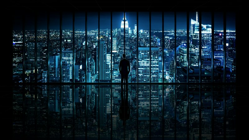 Silhouetted businessman standing in a high-rise office overlooking glowing Manhattan skyline at n...
