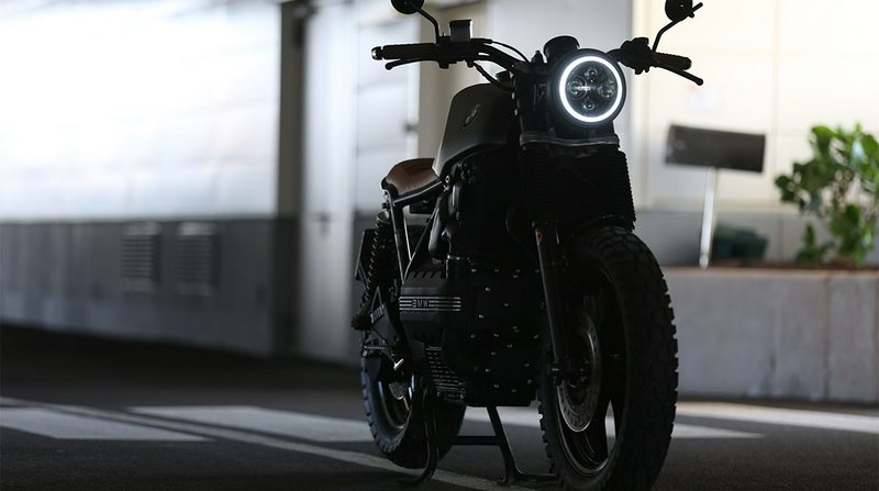 Custom matte black BMW scrambler motorcycle with glowing LED headlight parked in an indoor parkin...