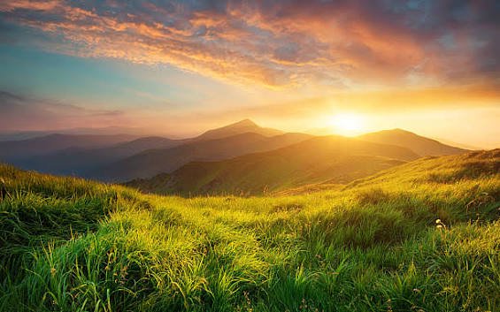 Golden sunset spilling over layered mountain ranges viewed from a lush grassy alpine hillside