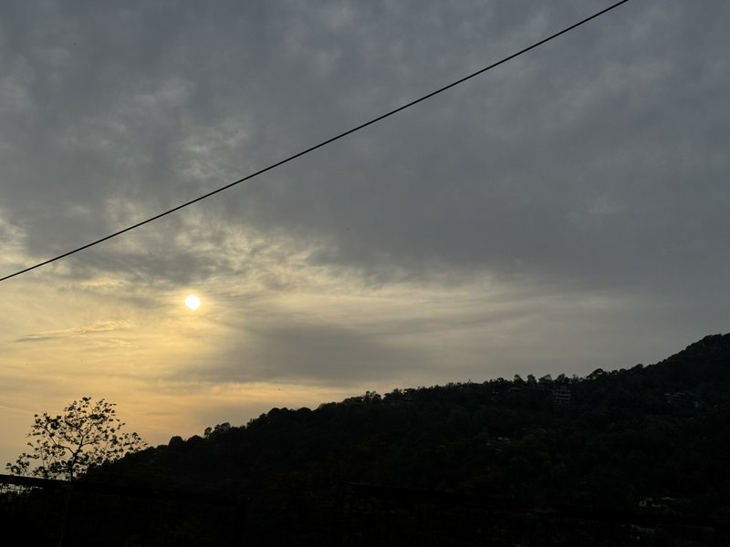 Low golden sun shines through cloudy dusk sky above dark silhouetted forested hill ridge