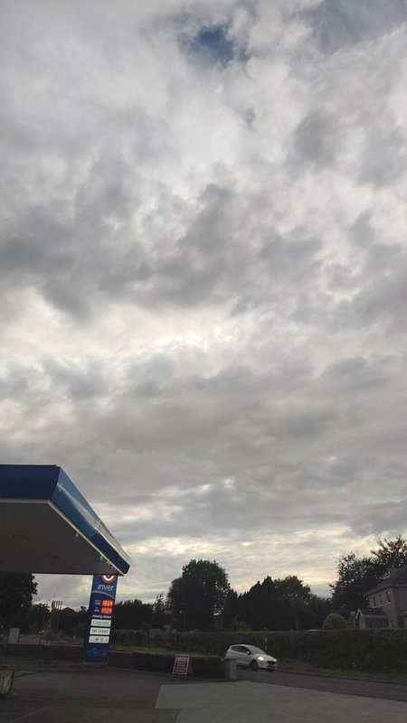 Overcast cloudy sky above a roadside Inver gas station, white car driving along adjacent suburban...
