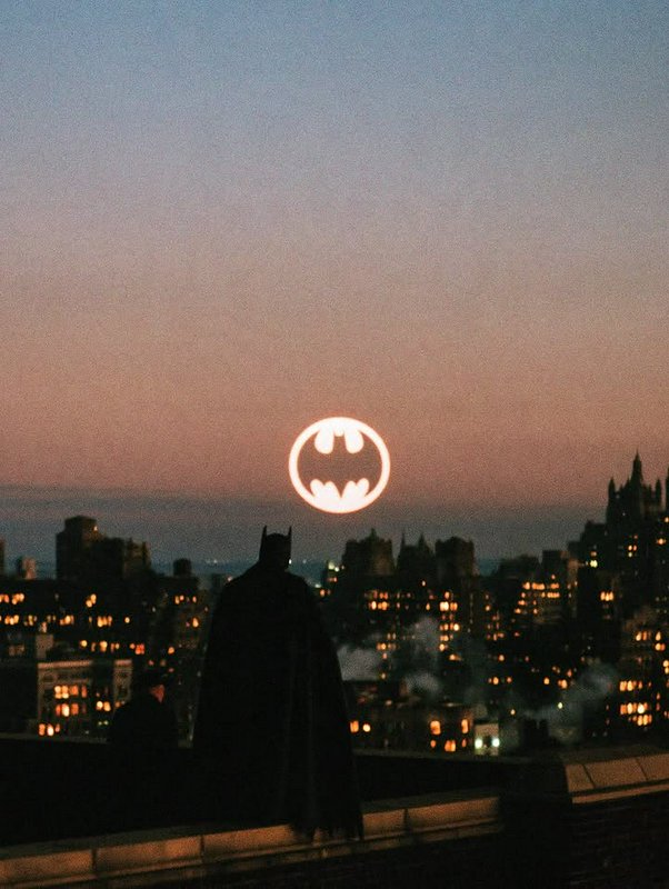 Silhouette of Batman standing on a Gotham rooftop facing the glowing Bat-signal at dusk