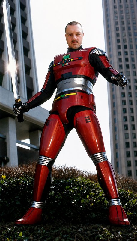 Cosplayer in glossy red tokusatsu armor holds a silver sword standing in front of downtown skyscr...