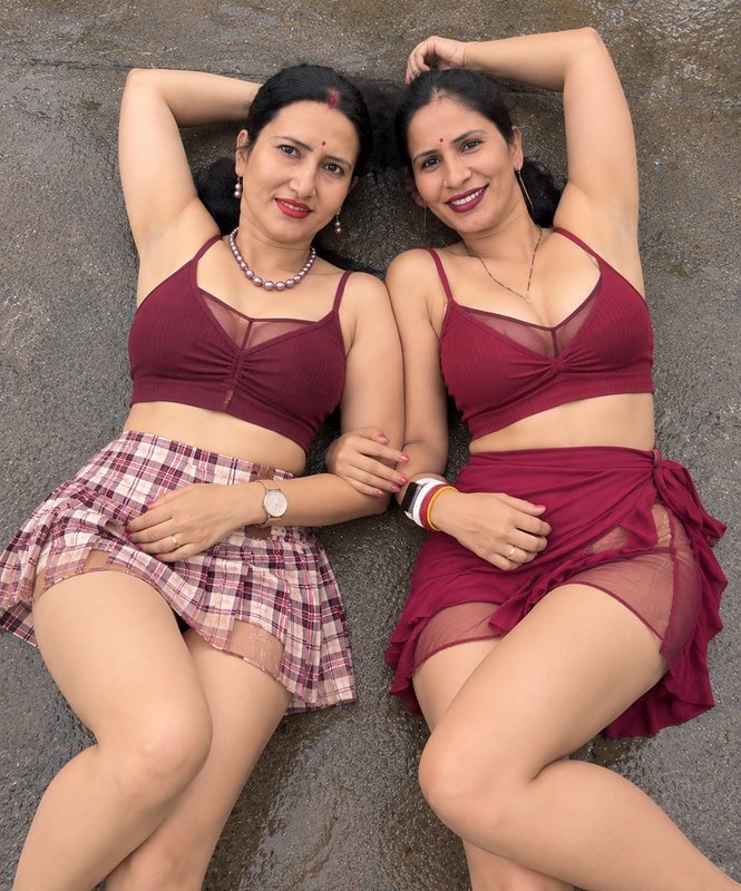 Two smiling South Asian women in matching maroon tops lie side-by-side on wet paved ground, posin...