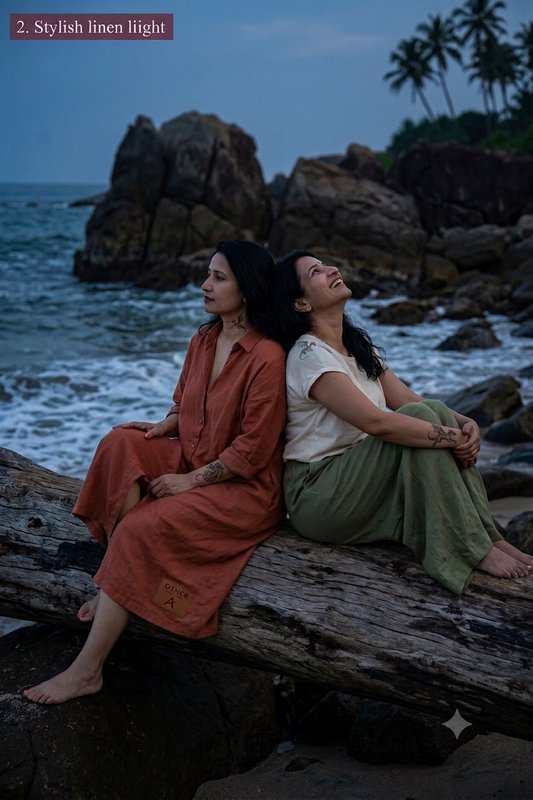 Two women sit back-to-back on driftwood at a rocky twilight shore, wearing casual earth-toned lin...