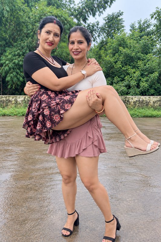 Two smiling South Asian women pose playfully, with one friend carrying the other on wet rain-soak...