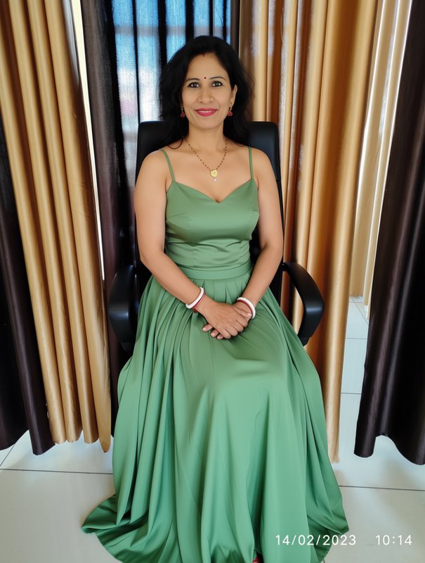 Smiling Indian woman in a flowing sage green satin gown seated in a black office chair indoors