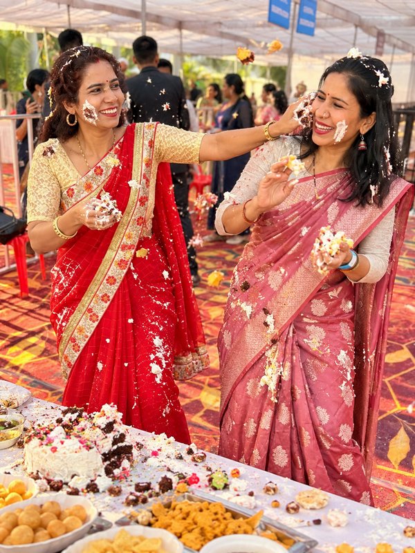 Two women in traditional sarees laugh as they playfully smear cake on each other at an outdoor ce...
