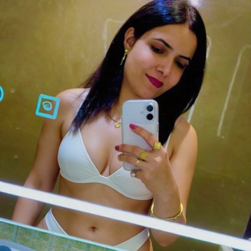 Woman in a white bikini takes a mirror selfie, holding a smartphone and wearing gold jewelry agai...