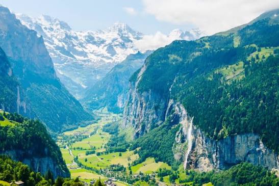 Sunlit panoramic view of alpine valley, cascading cliff waterfall, and snow-capped Swiss mountain peaks
