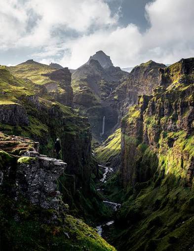 Lone hiker on a cliff edge overlooks a mossy Icelandic canyon with waterfall and jagged mountain ...
